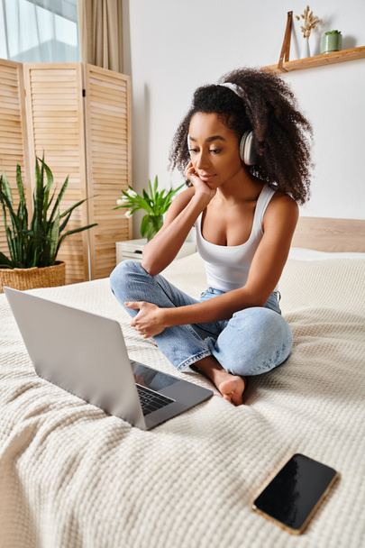 Una mujer afroamericana rizada se sienta en una cama, enfocada en la pantalla de su computadora portátil en un dormitorio moderno. - Foto, imagen