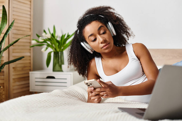 Μια σγουρή Αφρο-Αμερικανίδα γυναίκα σε ένα μπλουζάκι χαλαρώνει στο κρεβάτι, ακούγοντας μουσική στο τηλέφωνό της σε ένα μοντέρνο υπνοδωμάτιο. - Φωτογραφία, εικόνα