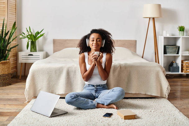 Μια σγουρή Αφρο-Αμερικανίδα με μπλουζάκι κάθεται γαλήνια στο πάτωμα μπροστά σε ένα μοντέρνο κρεβάτι.. - Φωτογραφία, εικόνα