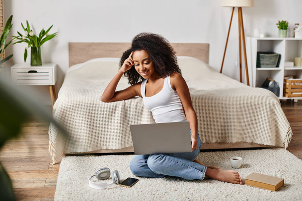 Una mujer afroamericana rizada en una camiseta sin mangas está sentada en el suelo, absorta en el uso de una computadora portátil en un dormitorio moderno. - Foto, imagen
