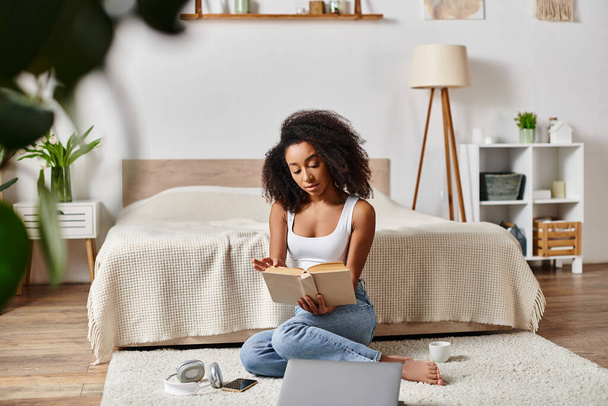 Μια σγουρή Αφρο-Αμερικανίδα με ένα μπλουζάκι που κάθεται στο πάτωμα, απορροφημένη στην ανάγνωση ενός βιβλίου σε μια μοντέρνα κρεβατοκάμαρα.. - Φωτογραφία, εικόνα