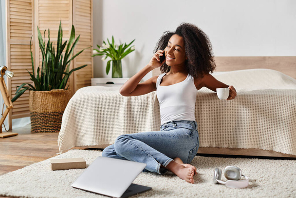 Askılı tişörtlü kıvırcık Afrikalı Amerikalı bir kadın yerde oturuyor, modern bir yatak odasında telefon görüşmesi yapıyor.. - Fotoğraf, Görsel