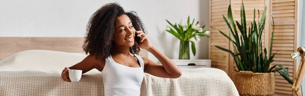 Kolsuz tişörtlü kıvırcık Afrikalı Amerikalı kadın modern yatakta otururken cep telefonuyla sohbet ediyor.. - Fotoğraf, Görsel