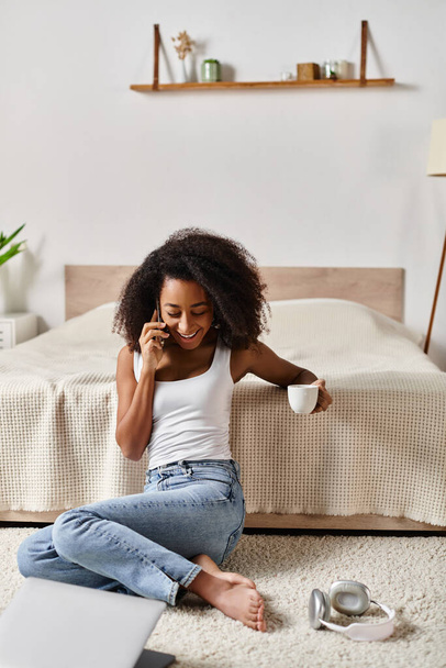 Femme afro-américaine bouclée en débardeur, assise sur le sol dans une chambre moderne, engagée dans une conversation sur un téléphone portable. - Photo, image