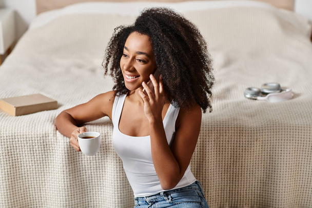 Curly African American γυναίκα σε δεξαμενή κορυφή απολαμβάνει μια στιγμή χαλάρωσης, πίνοντας καφέ σε ένα άνετο κρεβάτι σε ένα σύγχρονο υπνοδωμάτιο. - Φωτογραφία, εικόνα