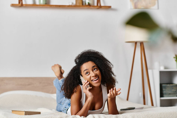 Μια σγουρή Αφρο-Αμερικανίδα με ένα μπλουζάκι είναι άνετα ξαπλωμένη σε ένα κρεβάτι, σε μια τηλεφωνική συνομιλία σε ένα μοντέρνο υπνοδωμάτιο.. - Φωτογραφία, εικόνα