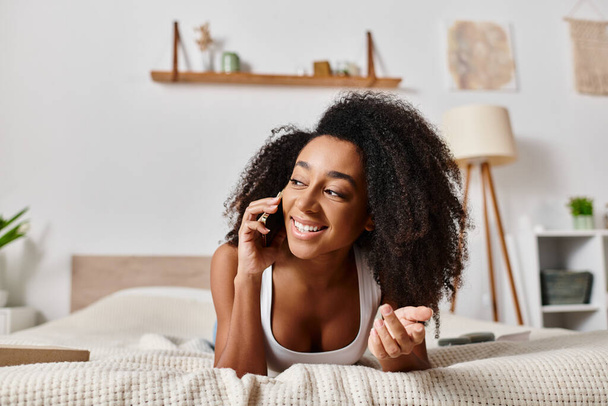 ベッドの上にラウンジングタンクのカーリーアフリカ系アメリカ人女性, 電話での会話に魅了. - 写真・画像