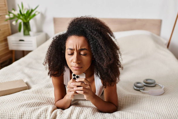 Curly Afrikkalainen Amerikkalainen nainen toppi, makaa sängyllä pitäen matkapuhelin modernissa makuuhuoneessa. - Valokuva, kuva