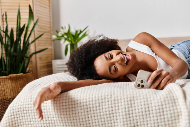 Curly afro-americano mulher no topo do tanque relaxante na cama, segurando um smartphone na mão. - Foto, Imagem