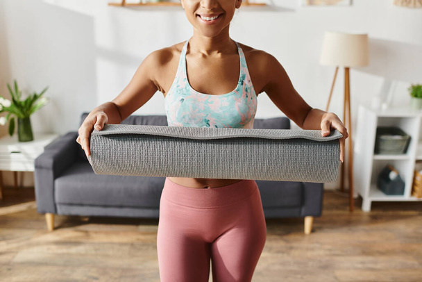 Mujer afroamericana en ropa deportiva sostiene una esterilla de yoga con una sonrisa brillante, que encarna bienestar y positividad. - Foto, imagen