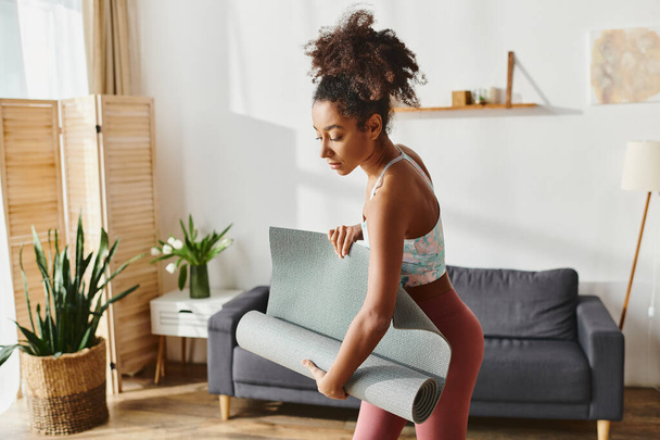 Curly Afro-Amerikaanse vrouw in actieve kleding houdt een yoga mat in een gezellige woonkamer, klaar voor een rustige training. - Foto, afbeelding