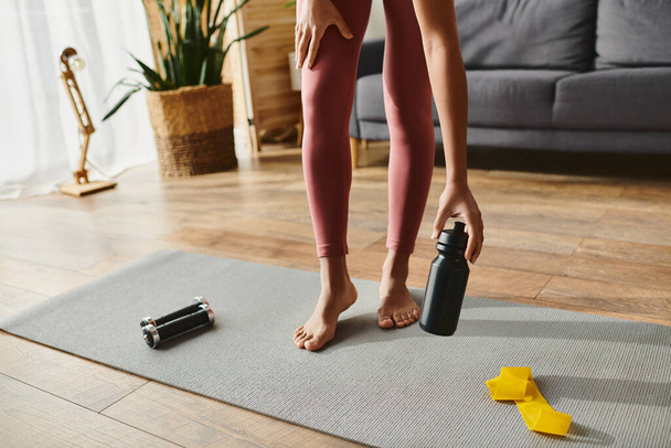 cropped African American woman in activewear standing on yoga mat, holding a bottle of water. - Photo, Image