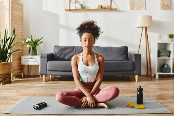 Curly mulher afro-americana em activewear praticando ioga em um tapete em um ambiente acolhedor sala de estar. - Foto, Imagem