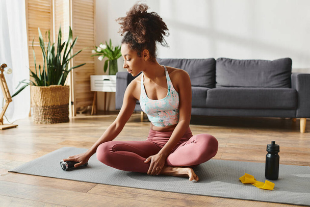 Кучерява афроамериканка в активному одязі, сидячи на килимку для йоги в затишній вітальні, практикуючи уважність. - Фото, зображення