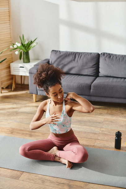 Кучерява афроамериканка в активному одязі практикує йогу на килимку в затишній обстановці вітальні. - Фото, зображення