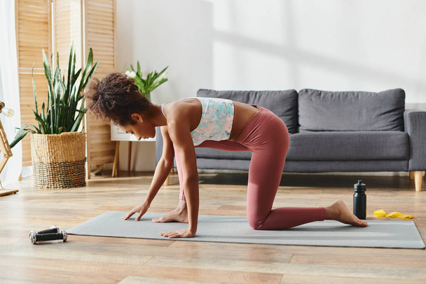 Una mujer afroamericana rizada en ropa deportiva equilibra elegantemente una esterilla de yoga en una pose desafiante de yoga. - Foto, imagen