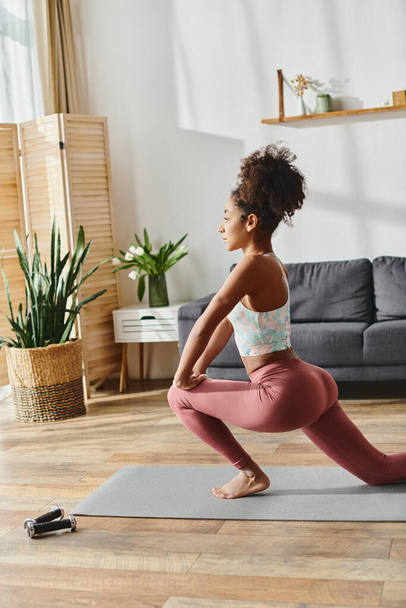 Kędzierzawa Afroamerykanka w stroju aktywnym ćwiczy jogę w swoim przytulnym salonie wypełnionym naturalnym światłem. - Zdjęcie, obraz