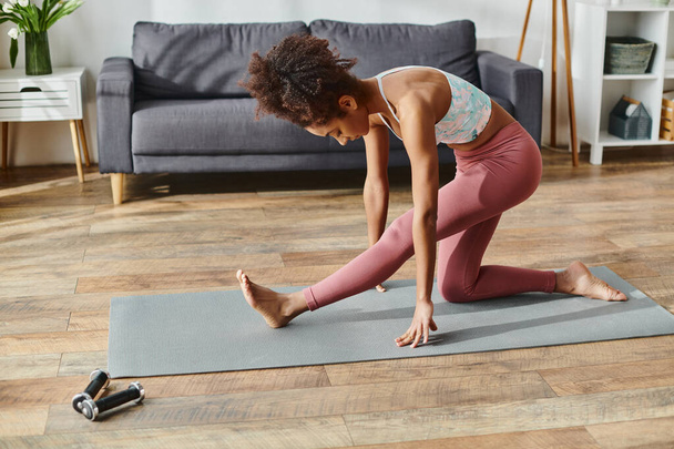 Una donna afroamericana riccia in activewear regge con grazia una posa yoga su un tappetino da yoga a casa. - Foto, immagini