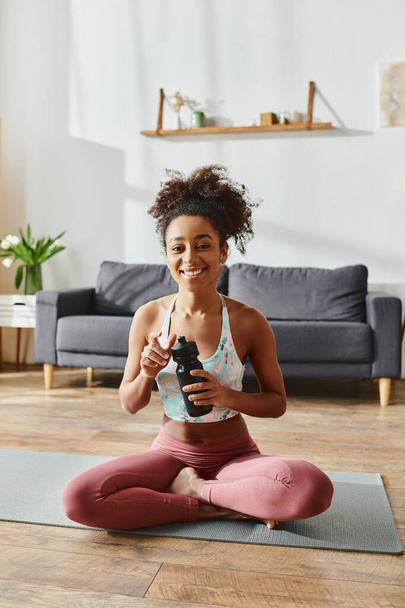 Een krullende Afro-Amerikaanse vrouw in activewear zit op een yoga mat, met een fles water. - Foto, afbeelding