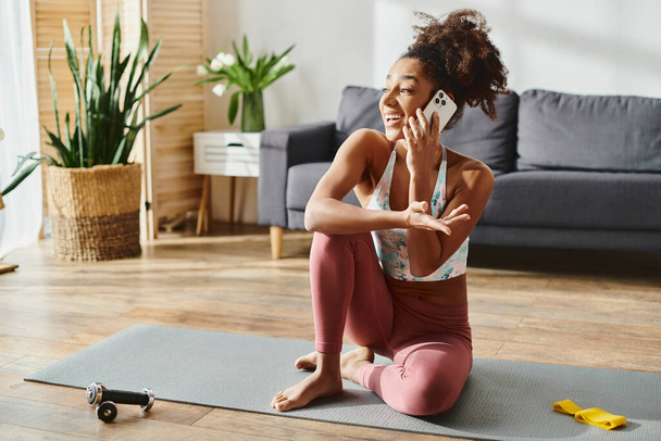 Kręcona Afroamerykanka w aktywnym ubraniu siedzi na macie do jogi, rozmawiając przez komórkę w domu. - Zdjęcie, obraz