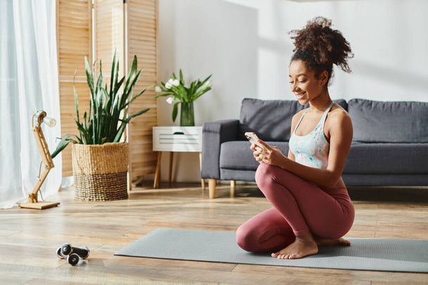 Donna afroamericana in activewear, seduta su tappetino yoga, focalizzata sullo schermo del telefono. - Foto, immagini