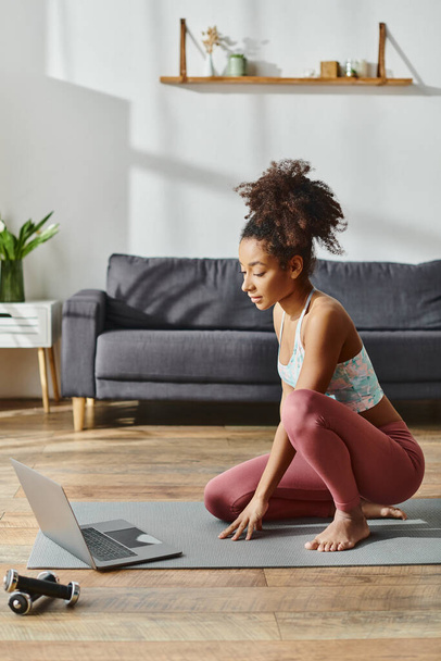Curly Afro-Amerikaanse vrouw in actieve dragen zit op yoga mat met laptop aan de voorkant, bezig met online fitness sessie. - Foto, afbeelding