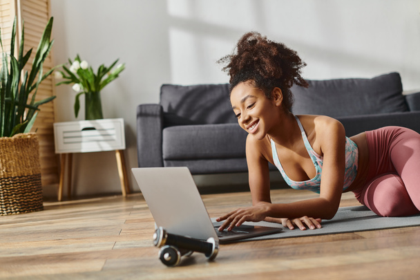 Una mujer afroamericana rizada en ropa activa, tendida en el suelo, se centra en su computadora portátil mientras hace ejercicio en casa. - Foto, imagen