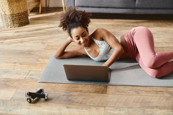Göndör afro-amerikai nő aktív ruhában jógázik egy szőnyegen, laptoppal. - Fotó, kép