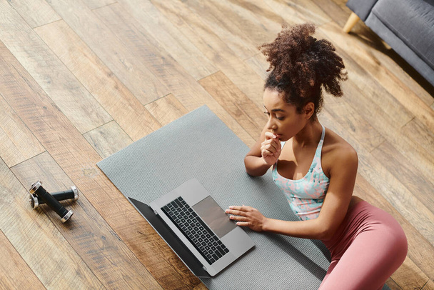 Ενεργό σγουρά Αφροαμερικανή γυναίκα πρακτική γιόγκα, ενώ πληκτρολογείτε σε φορητό υπολογιστή σε ένα στρώμα γιόγκα στο σπίτι. - Φωτογραφία, εικόνα