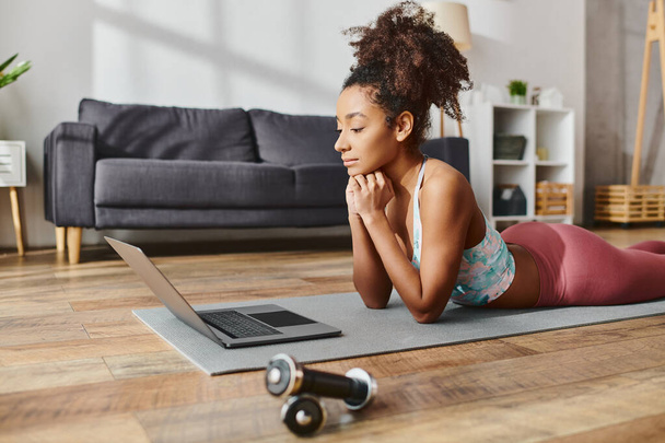 Eine lockige Afroamerikanerin in sportlicher Kleidung praktiziert Yoga, während sie zu Hause einen Laptop auf einer Matte benutzt. - Foto, Bild
