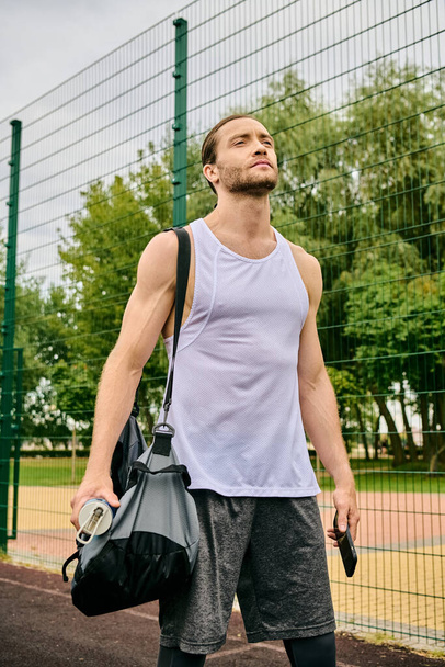 Ένας αποφασισμένος άντρας με αθλητικά περπατάει στο γήπεδο του τένις κρατώντας μια τσάντα. - Φωτογραφία, εικόνα
