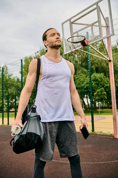 Een vastberaden man in sportkleding staat op een basketbalveld met een tas, klaar voor zijn volgende trainingssessie - Foto, afbeelding