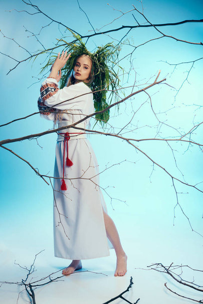 Μια νεαρή γυναίκα με λευκό φόρεμα και παραδοσιακή ενδυμασία στέκεται με χάρη μπροστά σε ένα μυστικιστικό δέντρο σε ένα σκηνικό στούντιο. - Φωτογραφία, εικόνα
