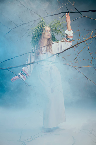 Μια νεαρή γυναίκα με λευκό φόρεμα στέκεται μέσα στην ομίχλη σε ένα μυστικιστικό δάσος, αποπνέοντας έναν αέρα αιθέριας ομορφιάς και μυστηρίου.. - Φωτογραφία, εικόνα