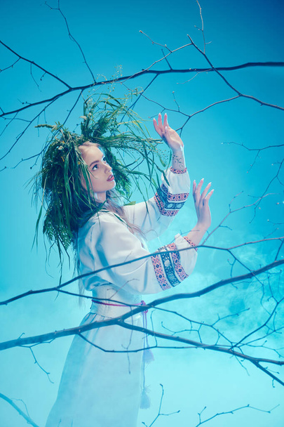 Μια νεαρή mavka στην παραδοσιακή ενδυμασία με περίτεχνα dreadlocks στέκεται με χάρη μπροστά από ένα μαγευτικό δέντρο. - Φωτογραφία, εικόνα