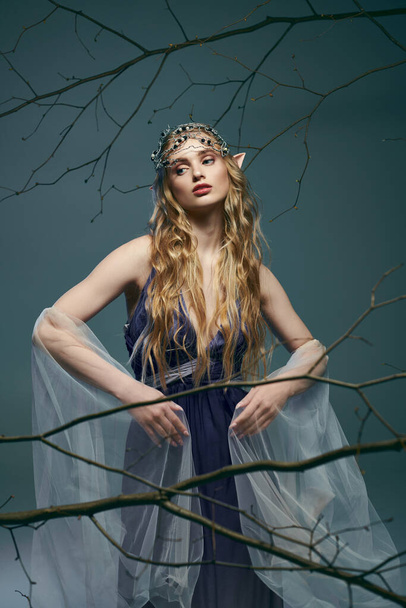幻想的なスタジオの設定で雄大な木の前に優雅に立っている美しい青いドレスの妖精のような若い女性. - 写真・画像