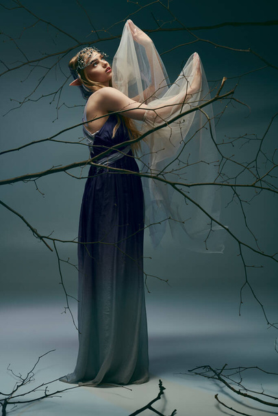 Młoda kobieta w długiej sukience delikatnie trzyma welon, emanując wróżkową aurą w studio. - Zdjęcie, obraz
