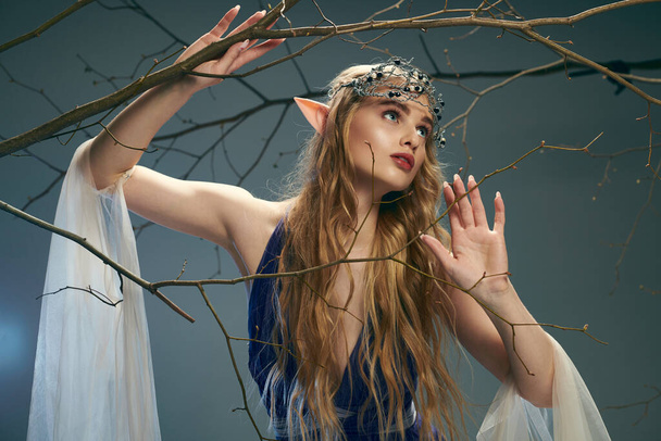流れる青いドレスを着ている若い女性は,魔法のファンタジーの設定で木の隣に優雅に立っています. - 写真・画像