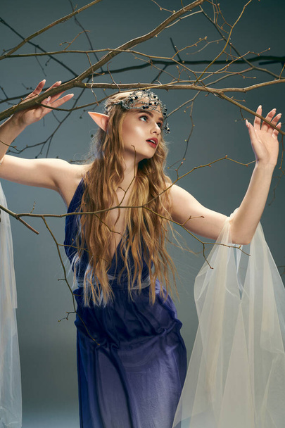 Μια νεαρή γυναίκα σε ένα μπλε φόρεμα ευγενικά κρατώντας ένα κλαδί δέντρου, ενσωματώνοντας την ουσία μιας νεράιδας πριγκίπισσας σε ένα μυστικιστικό σκηνικό. - Φωτογραφία, εικόνα
