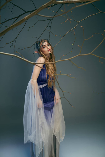 Μια νεαρή γυναίκα σε ένα μπλε φόρεμα στέκεται με χάρη δίπλα σε ένα δέντρο σε ένα παραμυθένιο και φανταστικό σκηνικό. - Φωτογραφία, εικόνα