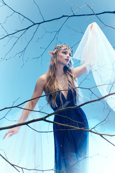 Μια νεαρή γυναίκα με ένα κυανό φόρεμα, που μοιάζει με μια πριγκίπισσα ξωτικό, στέκεται με χάρη πάνω σε ένα δέντρο με έναν παιχνιδιάρικο και παραμυθένιο τρόπο. - Φωτογραφία, εικόνα