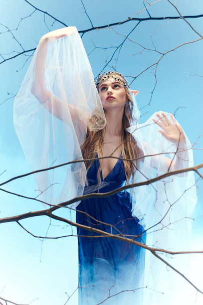 Mavi elbiseli genç bir kadın zarif bir şekilde beyaz bir duvak tutuyor, mistik bir stüdyoda peri gibi bir aura yayıyor.. - Fotoğraf, Görsel
