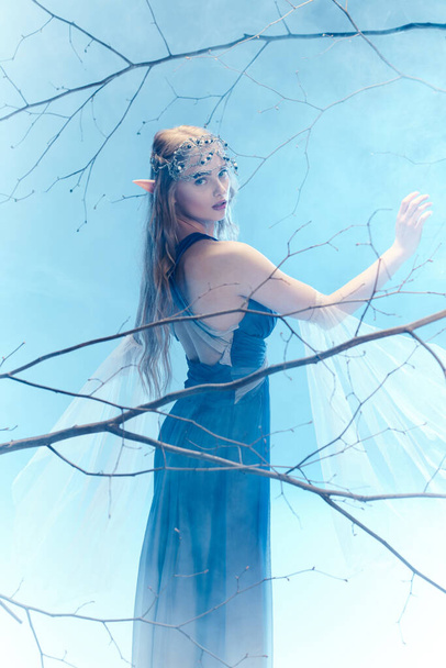 Mavi elbiseli genç bir kadın zarif bir şekilde bir ağaçta duruyor, mistik bir ormanda bir peri prensesinin özünü temsil ediyor.. - Fotoğraf, Görsel