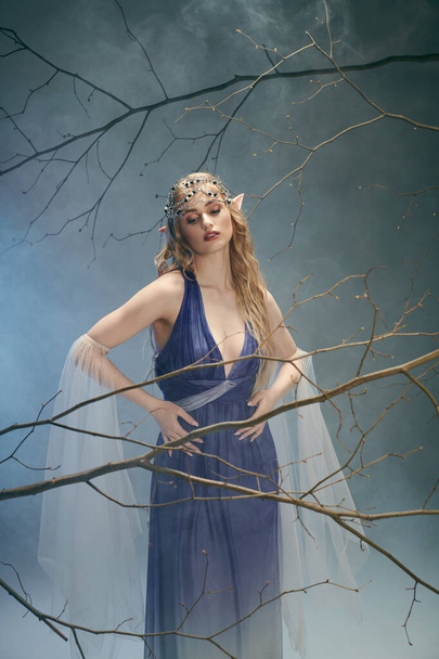 Μια νεαρή γυναίκα με μπλε φόρεμα στέκεται με χάρη μπροστά σε ένα μεγαλοπρεπές δέντρο, ενσαρκώνοντας μια παραμυθένια παρουσία σε ένα σκηνικό στούντιο.. - Φωτογραφία, εικόνα