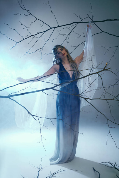 Egy fiatal nő, aki egy tündérhercegnőre hasonlít, elegánsan áll egy kék ruhában egy fenséges fa előtt.. - Fotó, kép