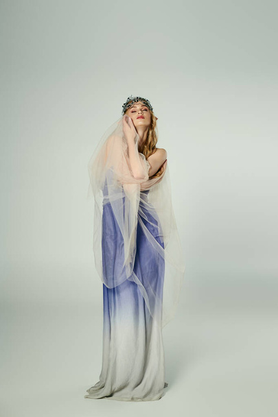Μια νεαρή γυναίκα ντυμένη με ένα μπλε και άσπρο φόρεμα με ένα πέπλο που κυλάει, ενσωματώνοντας την ουσία μιας νεράιδας πριγκίπισσας σε ένα σκηνικό στούντιο.. - Φωτογραφία, εικόνα