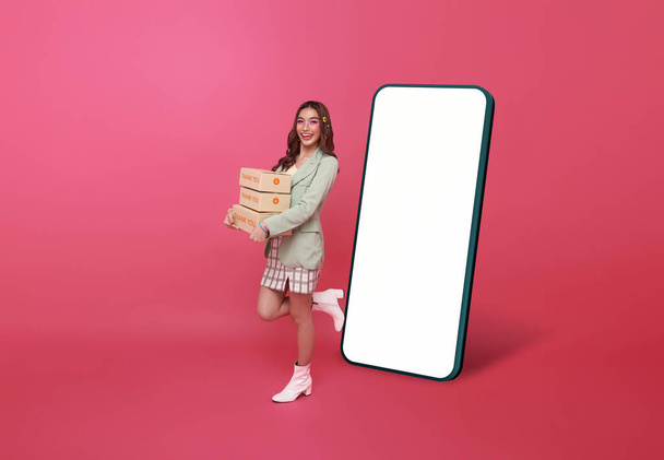 Heureuse femme asiatique souriant et tenant paquet boîte de colis avec maquette grand smartphone écran blanc vide isolé sur fond rose. - Photo, image