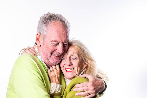 Esta imagen conmovedora cuenta con una pareja de ancianos caucásicos en un abrazo alegre. El hombre tiene el pelo gris y lleva un suéter verde claro, mientras que la mujer, con su pelo rubio, está en una aceituna suave - Foto, Imagen