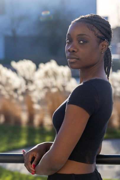 En esta imagen, una mujer africana pensativa se apoya en una barandilla de metal con un parque urbano de enfoque suave en el fondo. Ella mira a la distancia, su rostro parcialmente iluminado por la suave luz del sol, que - Foto, imagen