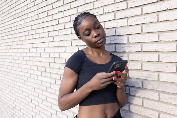 This image features a young African woman engrossed in her smartphone while standing against a white brick wall. Her posture is relaxed, and she holds the phone with both hands, indicating she is - Photo, Image